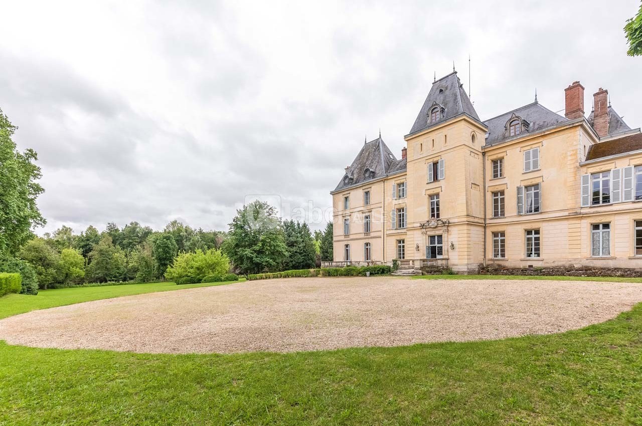 Façade d'un château en pierre claire entouré de pelouses et d'arbres sous un ciel nuageux.