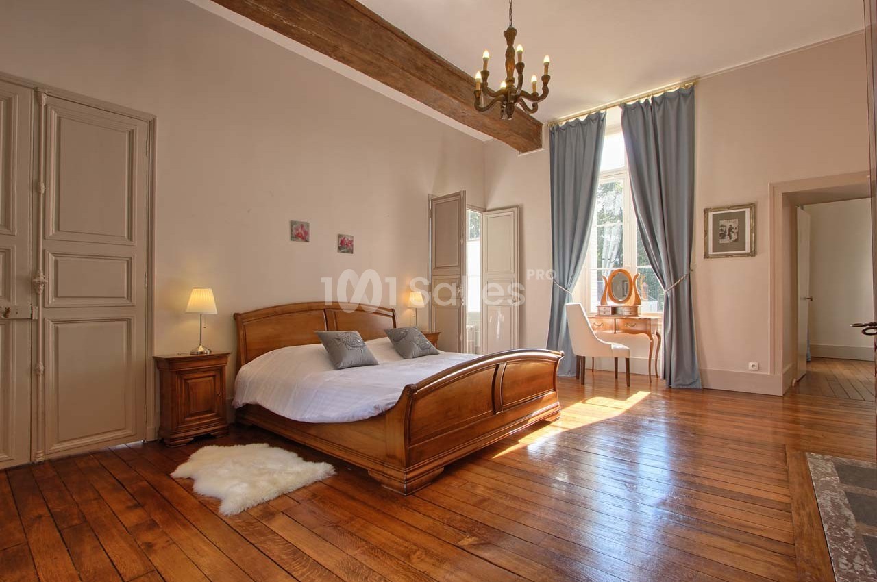 Chambre lumineuse avec lit double en bois, parquet, poutre apparente, bureau près d'une fenêtre et rideaux bleus.