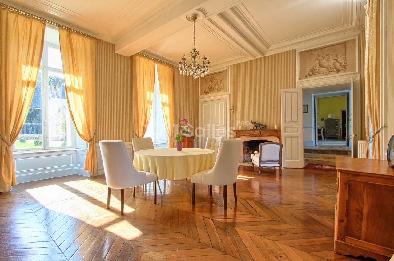 Salle à manger lumineuse avec parquet en bois, table ronde, chaises modernes, rideaux dorés et lustre élégant.