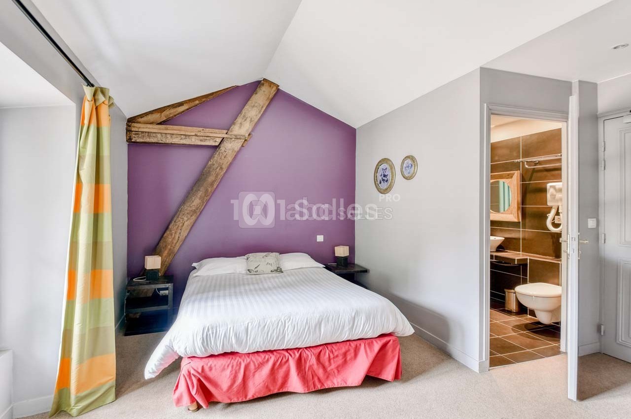 Chambre lumineuse avec lit double, mur violet avec poutre apparente, et salle de bain attenante visible à droite.