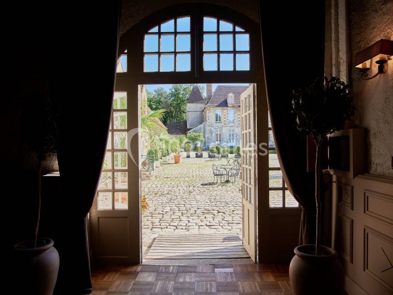 Vue depuis une porte ouverte sur une cour pavée ensoleillée avec des tables, des plantes et un bâtiment en pierre.