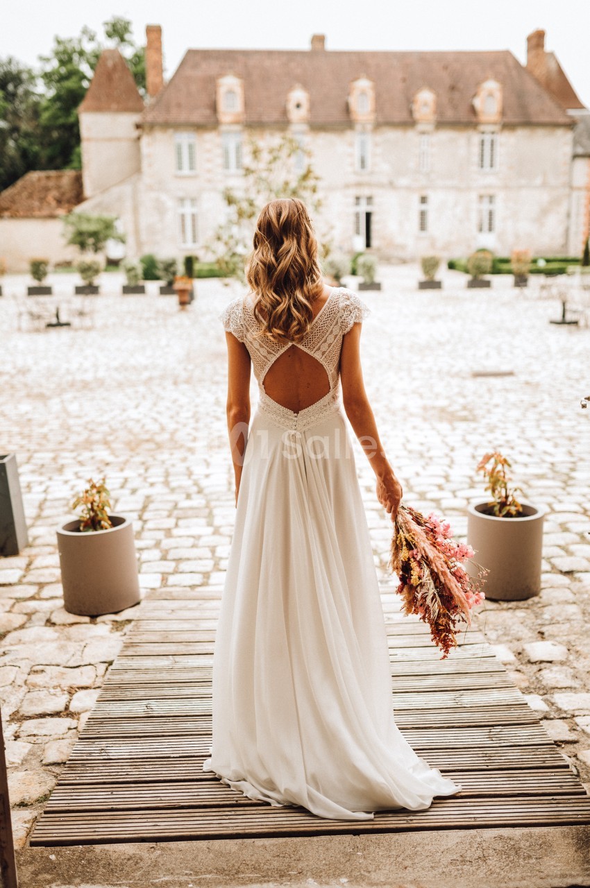 Une femme en robe de mariée blanche, vue de dos, tenant un bouquet de fleurs séchées devant un bâtiment en pierre.