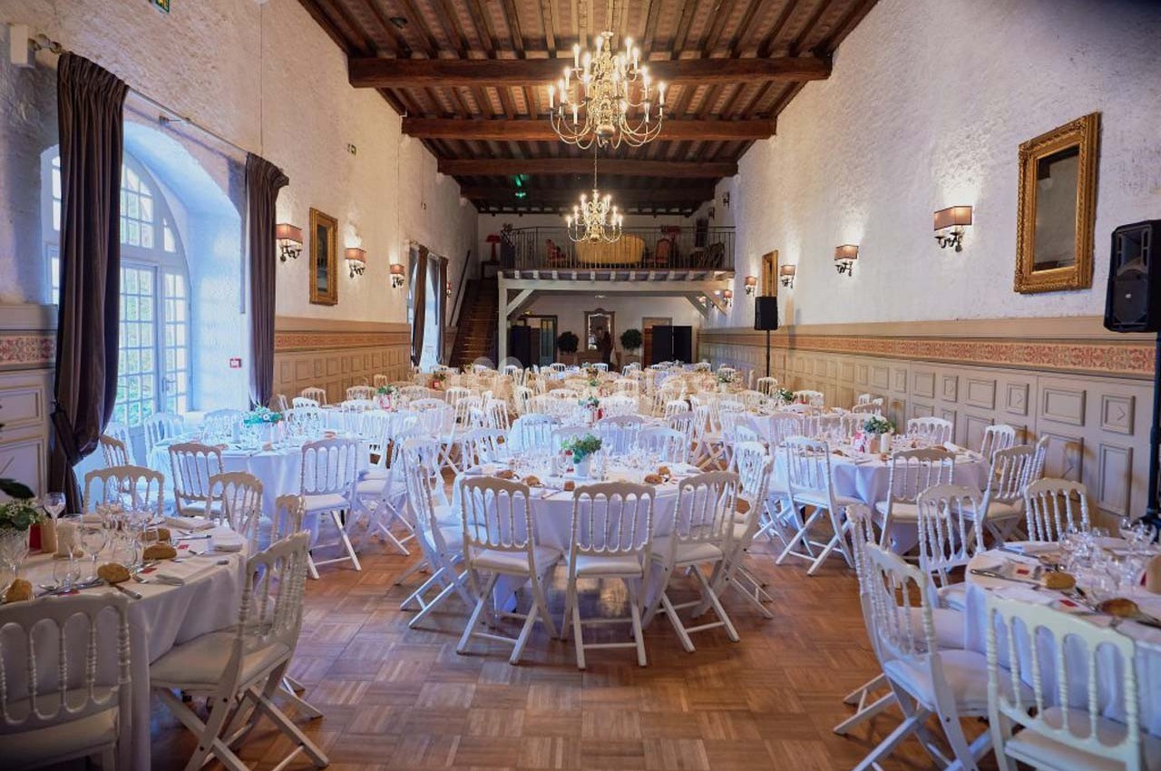 Salle de réception élégante avec tables rondes dressées, chaises blanches et lustres suspendus sous un plafond en bois.