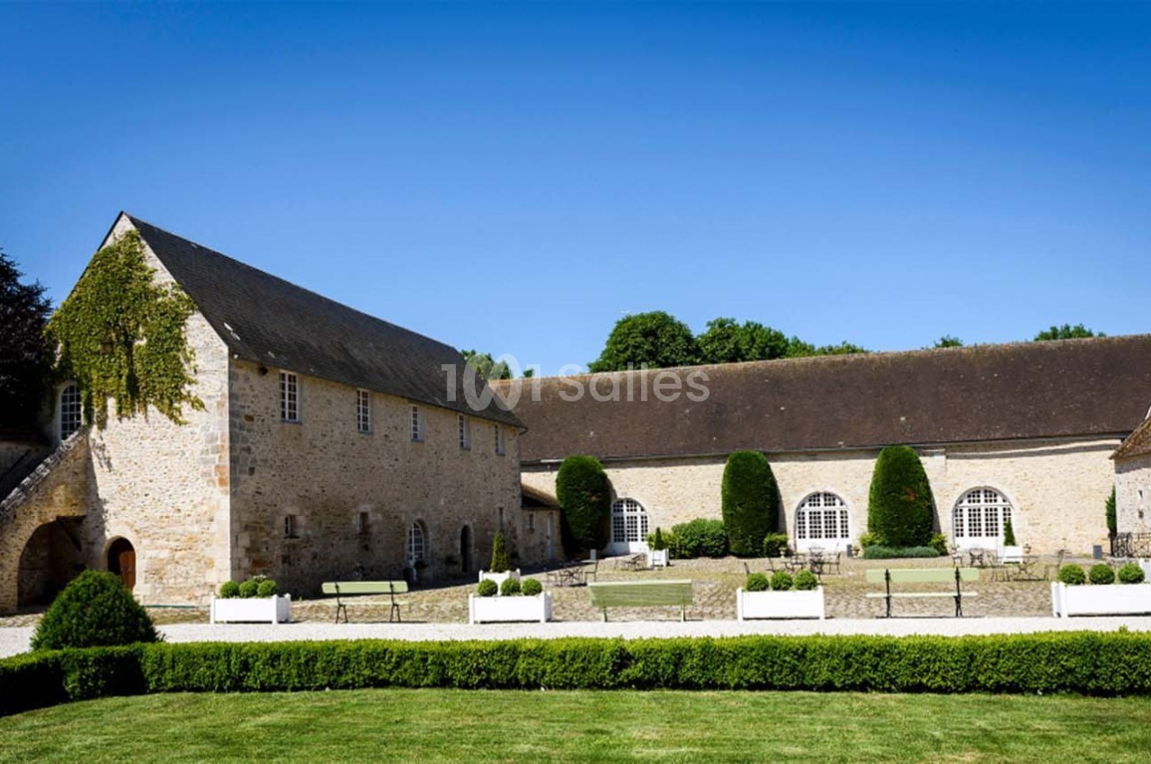 Bâtiments en pierre avec toits en ardoise entourant une cour pavée, bordée de haies et de bancs.