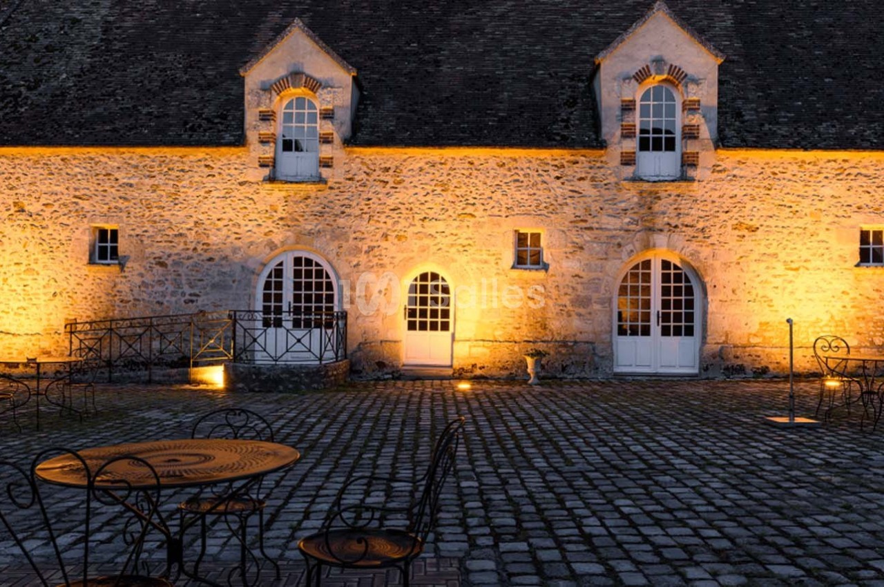 Façade en pierre d'un bâtiment ancien éclairé la nuit, avec tables et chaises en fer forgé sur une cour pavée.