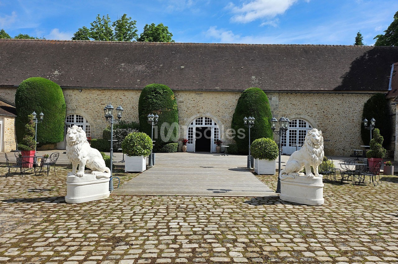Cour pavée avec deux statues de lions en pierre encadrant une entrée voûtée d'un bâtiment en pierre, entouré de verdure.