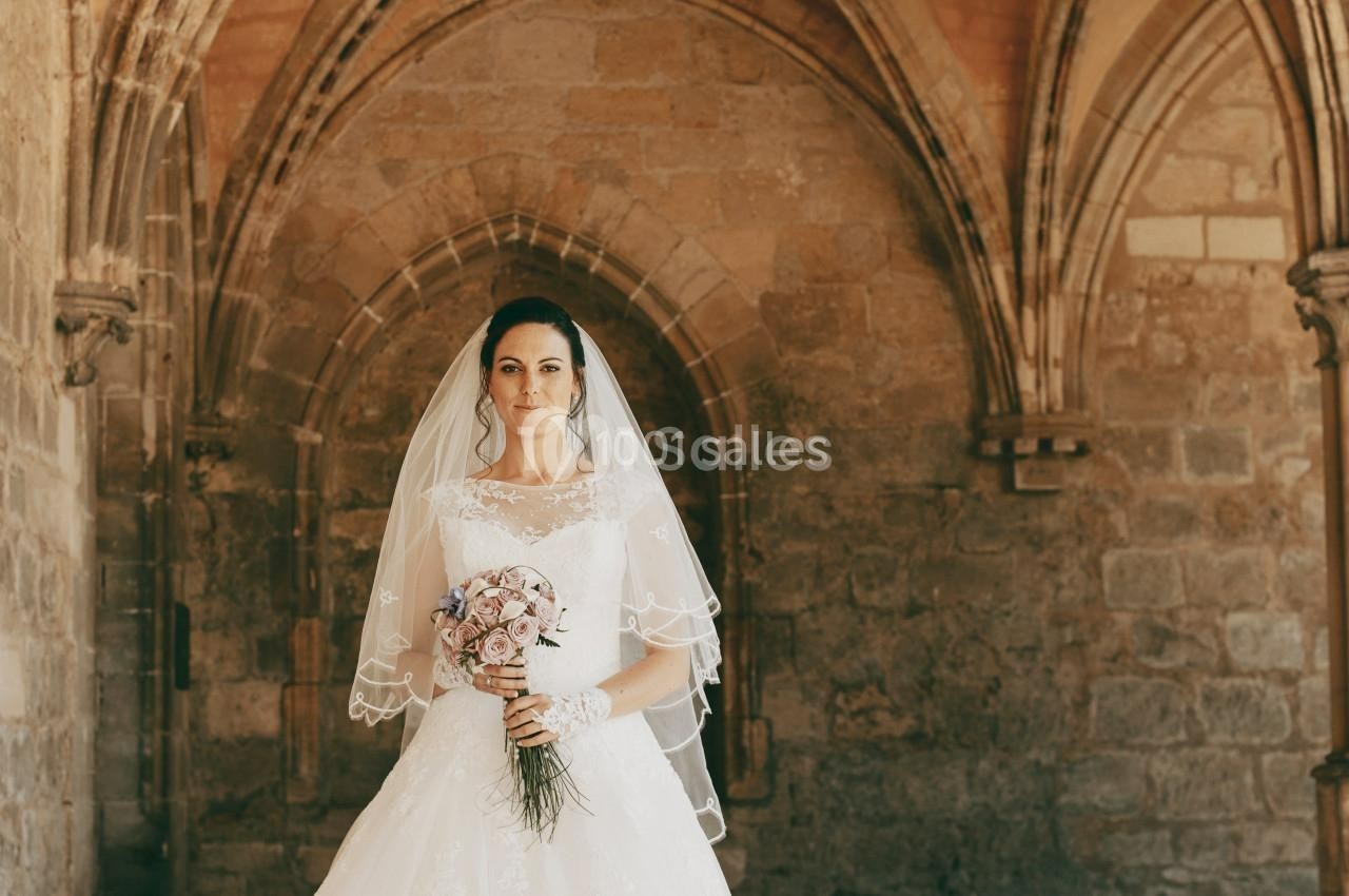 Une mariée en robe blanche et voile tient un bouquet de fleurs dans un cadre architectural en pierre ancienne.