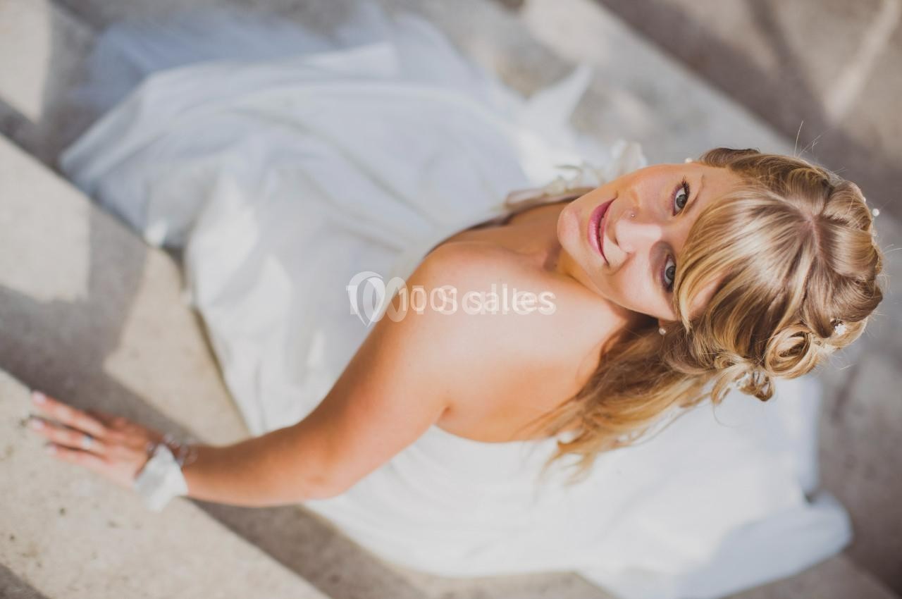 Femme en robe blanche assise sur des marches en pierre, regardant vers l'objectif avec une expression douce.