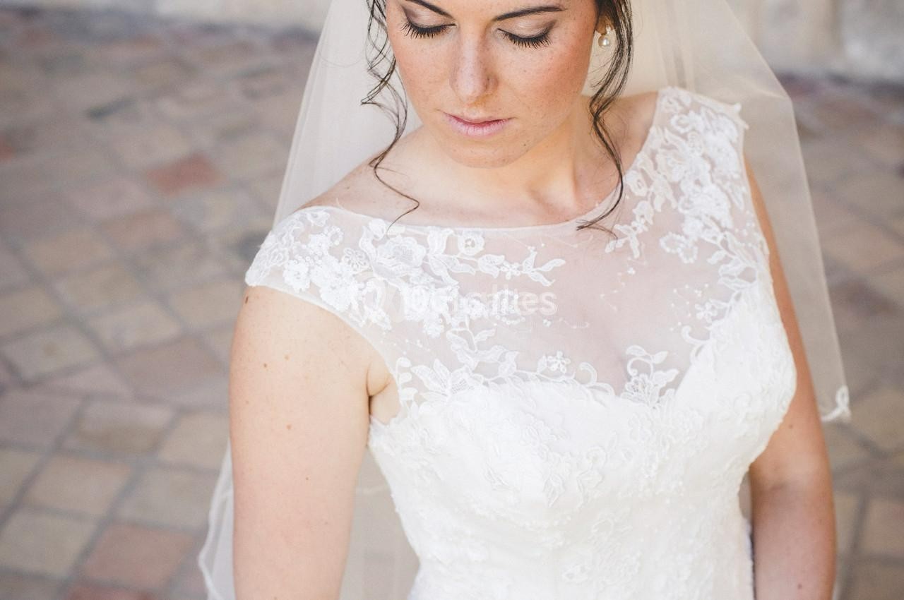 Femme en robe de mariée blanche avec un voile, regard baissé, sur un sol pavé.