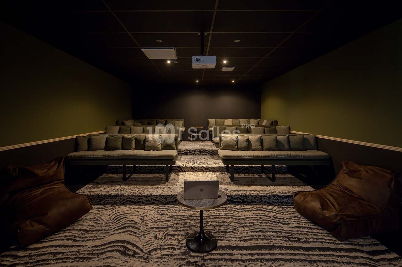 Salle de projection sombre avec plusieurs rangées de sièges rembourrés, poufs en cuir et un projecteur au plafond.