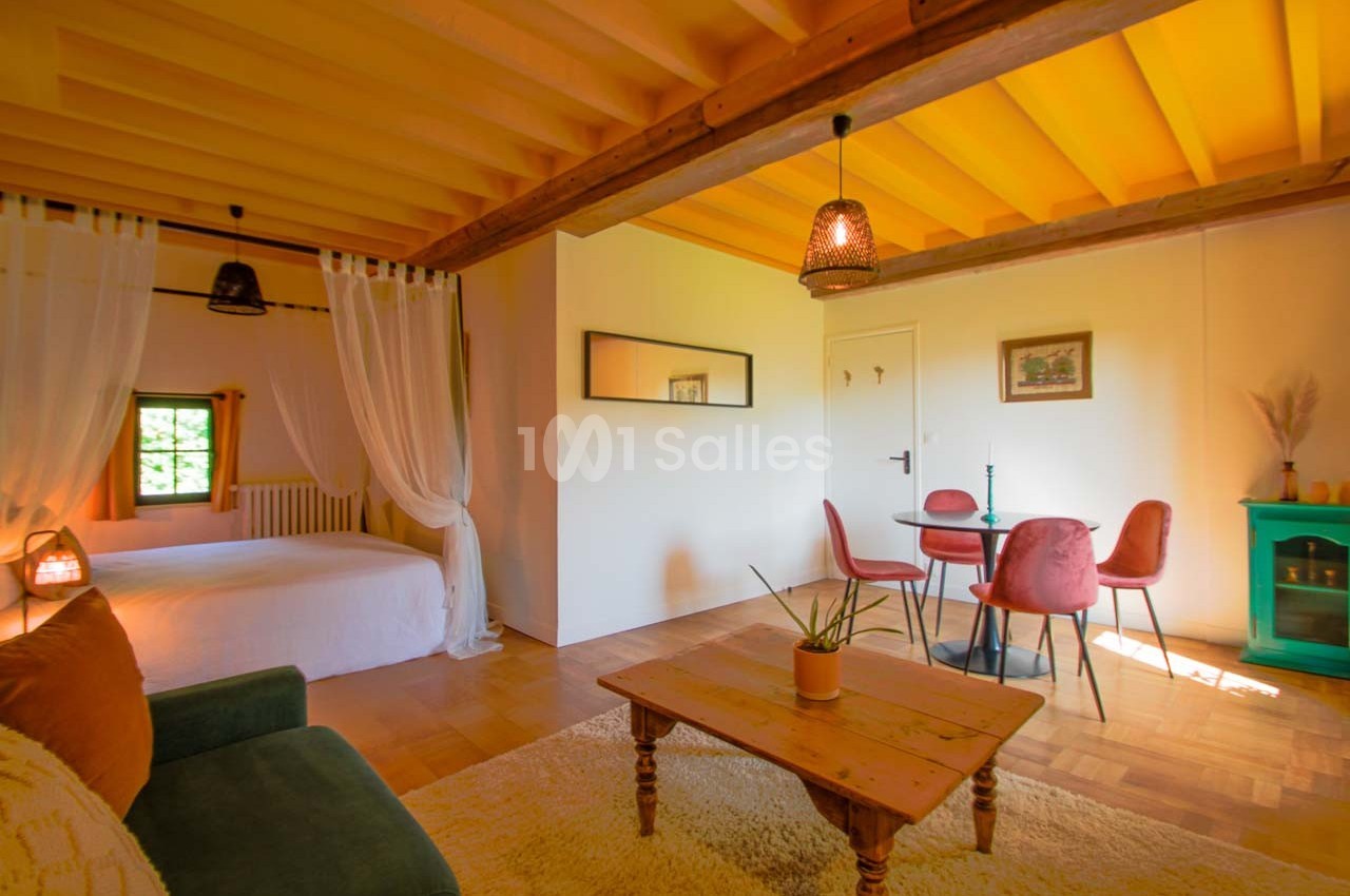 Salon lumineux avec canapé, table basse en bois, coin repas et lit séparé par un rideau, sous un plafond en bois jaune.