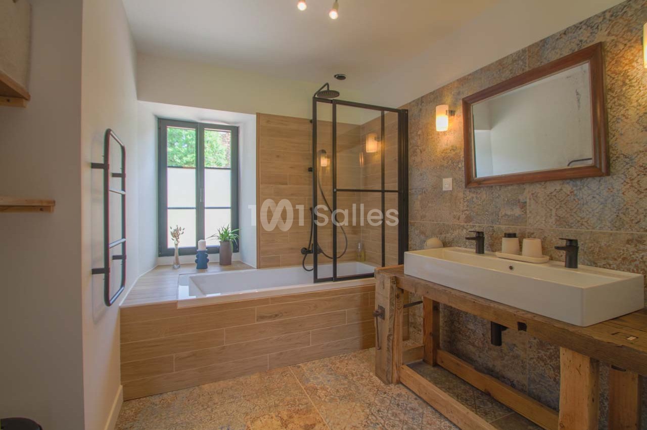 Salle de bain avec baignoire encastrée, paroi vitrée, lavabo double sur meuble en bois et mur en pierre naturelle.