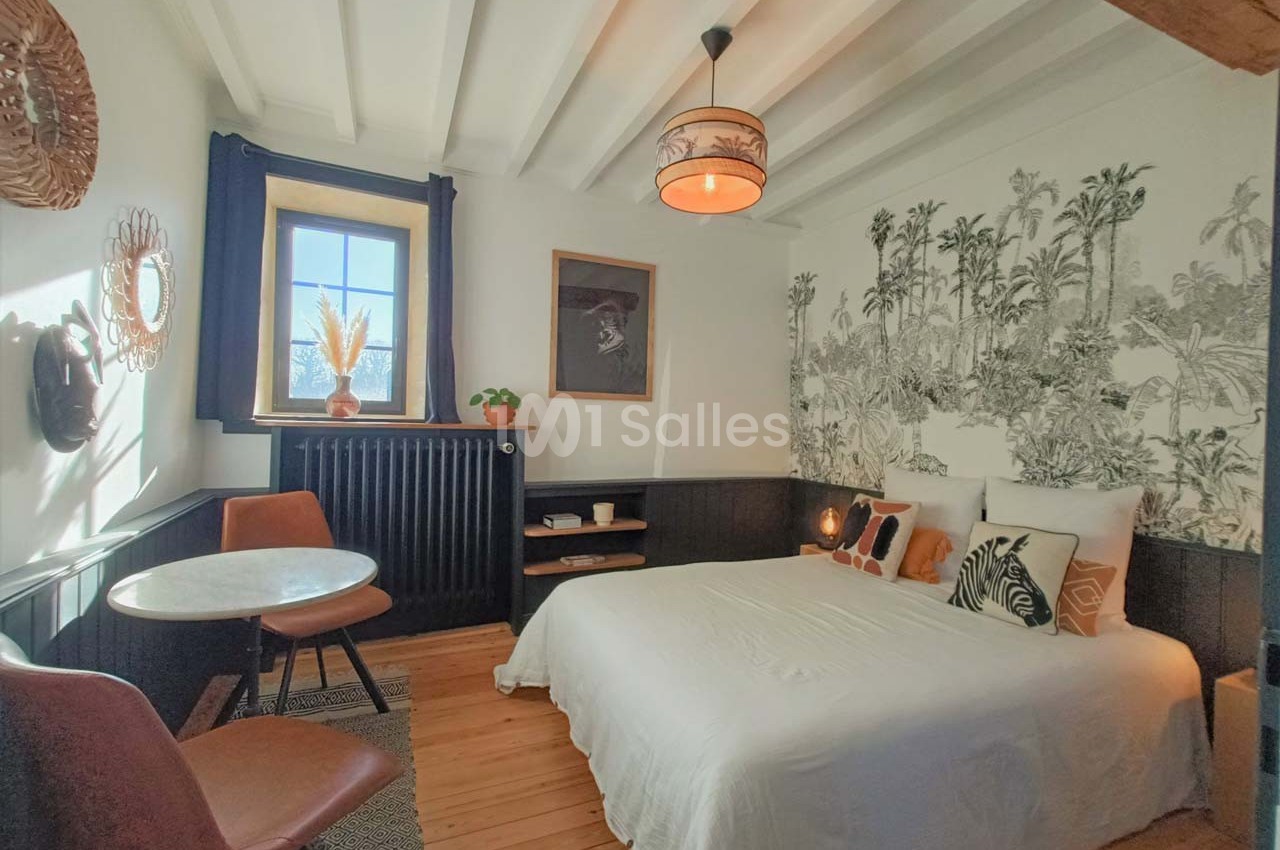 Chambre lumineuse avec lit double, table ronde, chaises en cuir et décoration murale tropicale en noir et blanc.