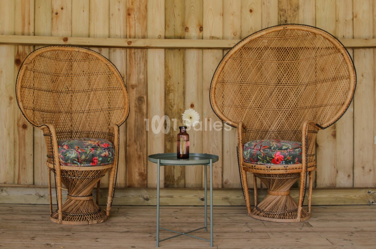 Deux fauteuils en rotin avec coussins fleuris encadrent une petite table métallique portant une bouteille et une fleur.