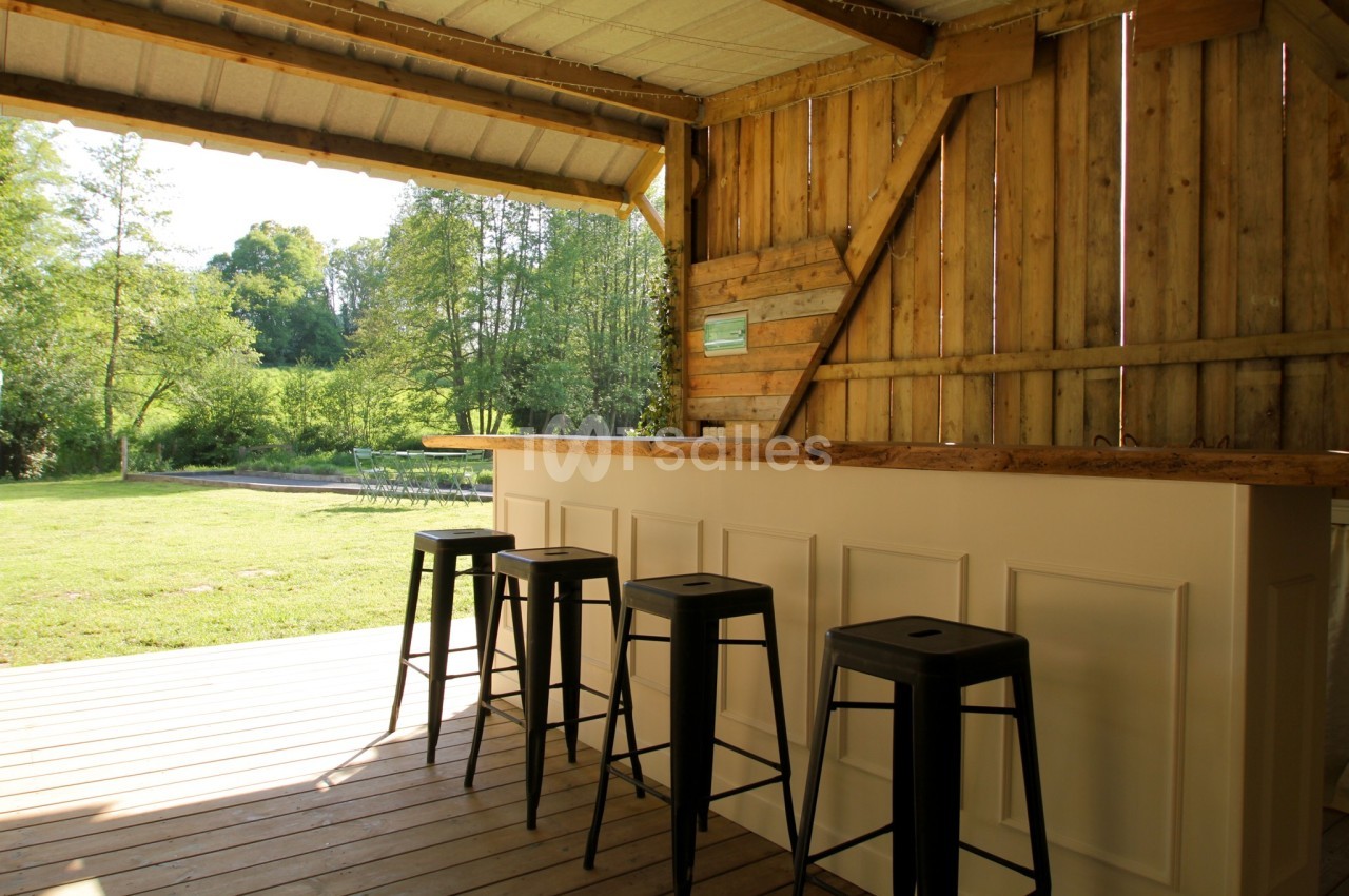 Bar en bois avec tabourets noirs sur une terrasse couverte, donnant sur un jardin verdoyant.
