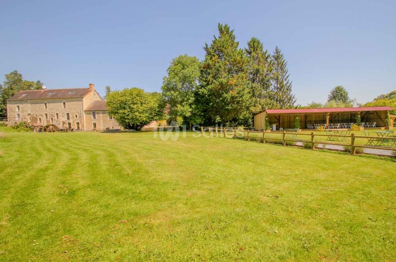 Vue d'un grand jardin verdoyant avec une maison en pierre à gauche et une structure couverte en bois à droite.