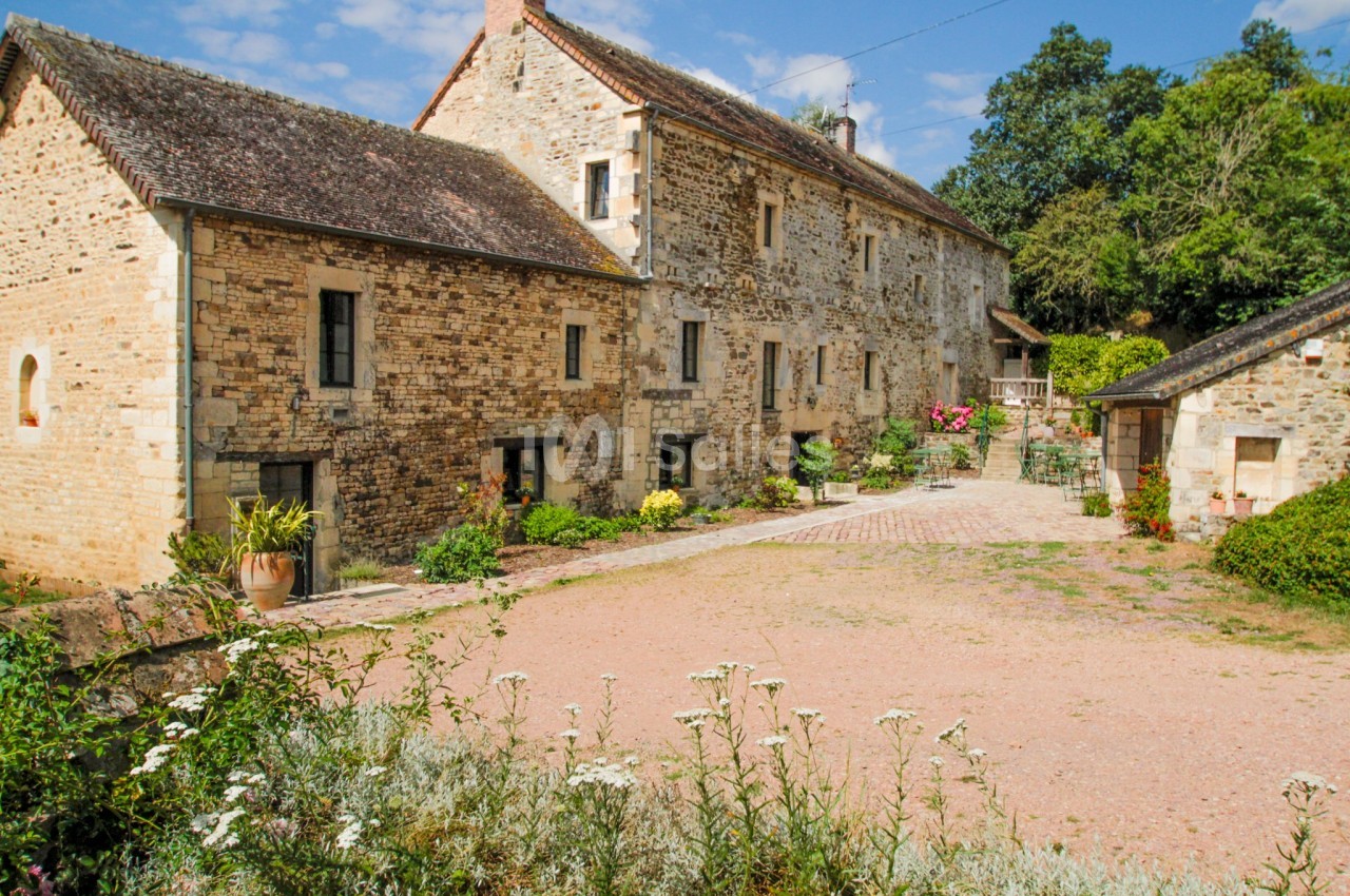Cour pavée devant une maison en pierre ancienne avec des volets en bois, entourée de végétation et fleurs.