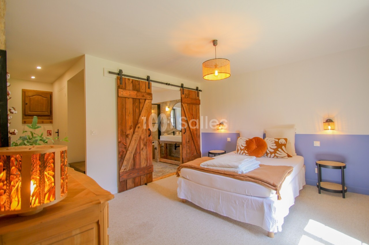 Chambre lumineuse avec lit double, portes coulissantes en bois, et salle de bain attenante visible en arrière-plan.