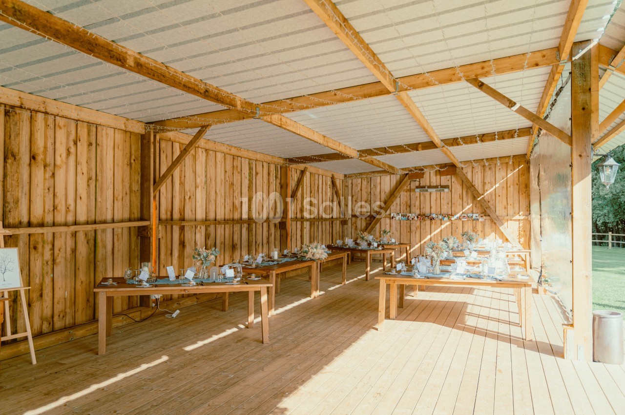 Salle en bois ouverte avec de grandes tables décorées pour un événement, éclairée par la lumière naturelle.