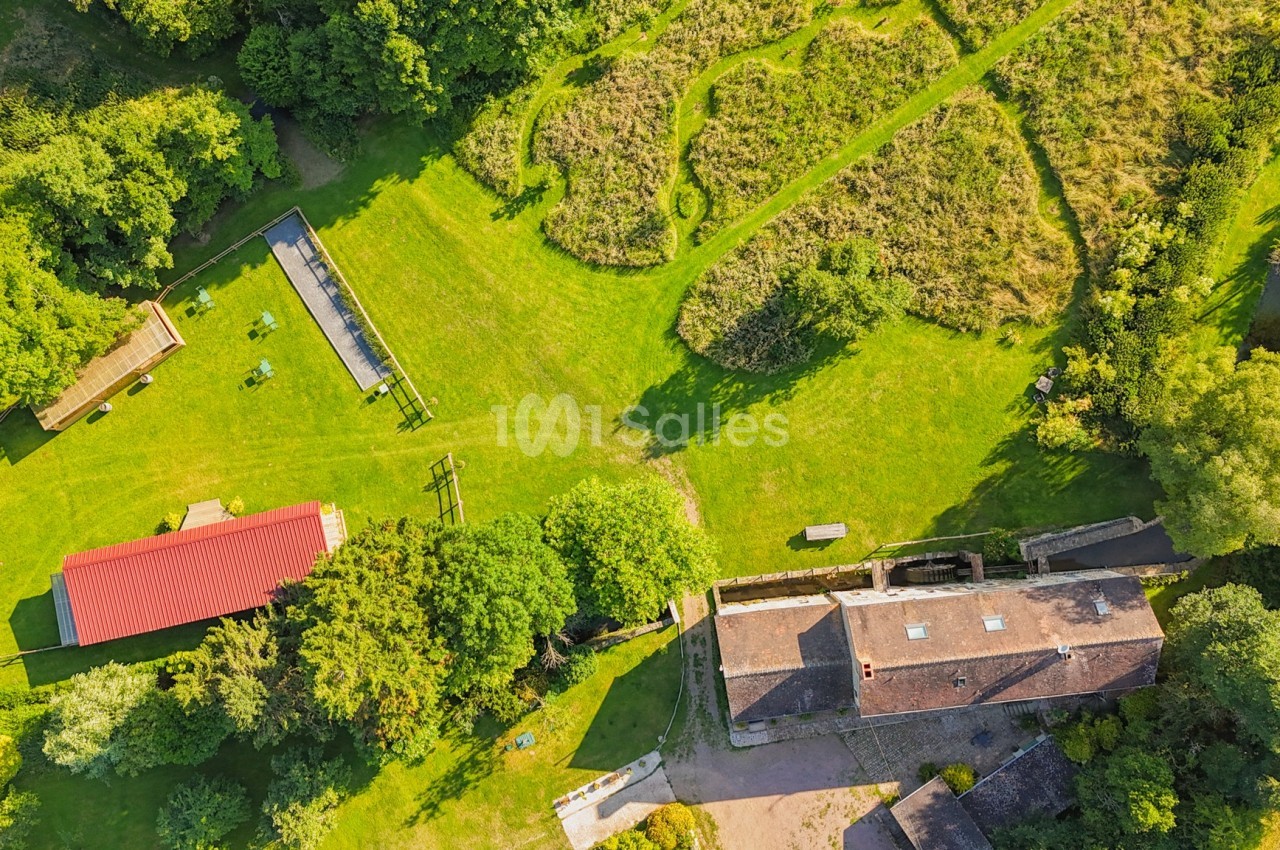 Vue aérienne d'une ferme avec bâtiments, espaces verts, enclos et chemins entourés de végétation dense.