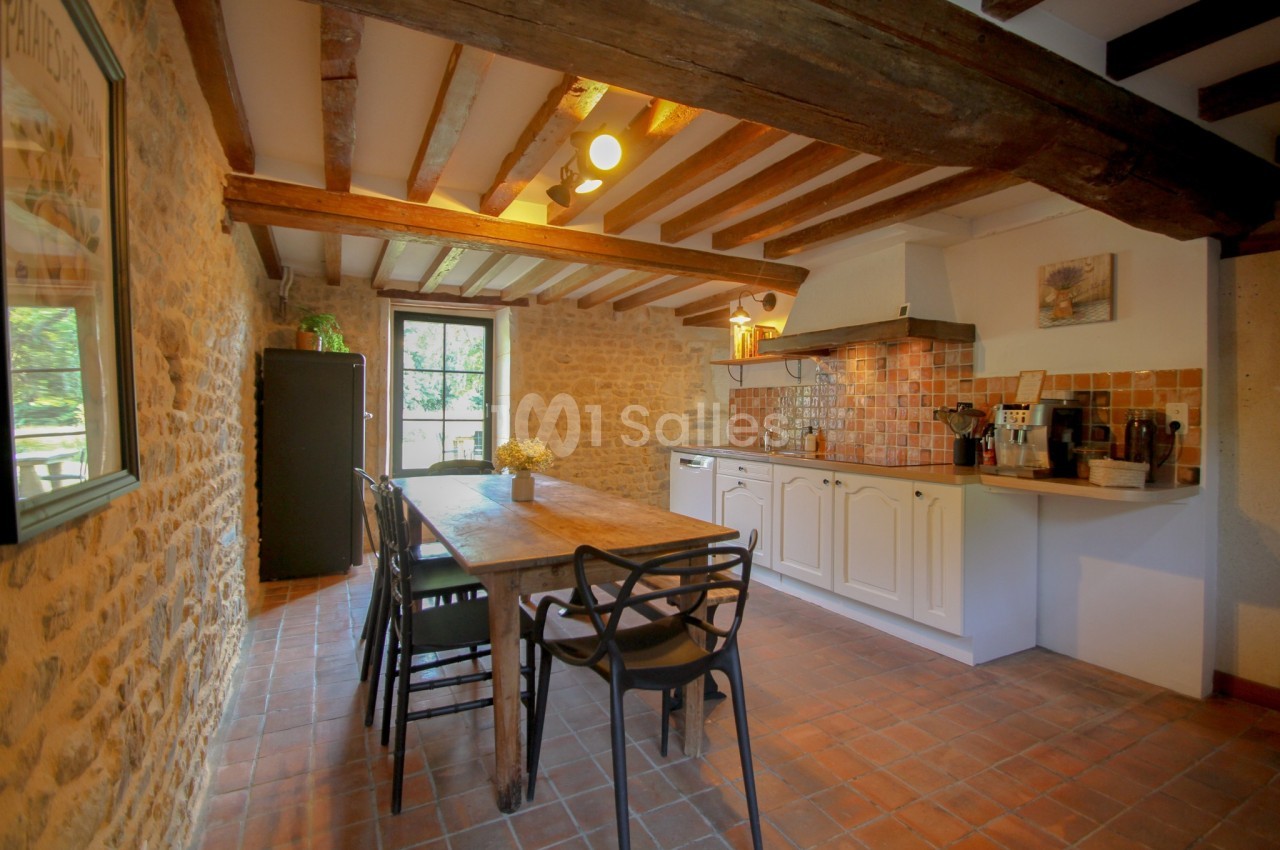 Cuisine rustique avec murs en pierre, poutres apparentes, table en bois, chaises noires et carrelage en terre cuite.