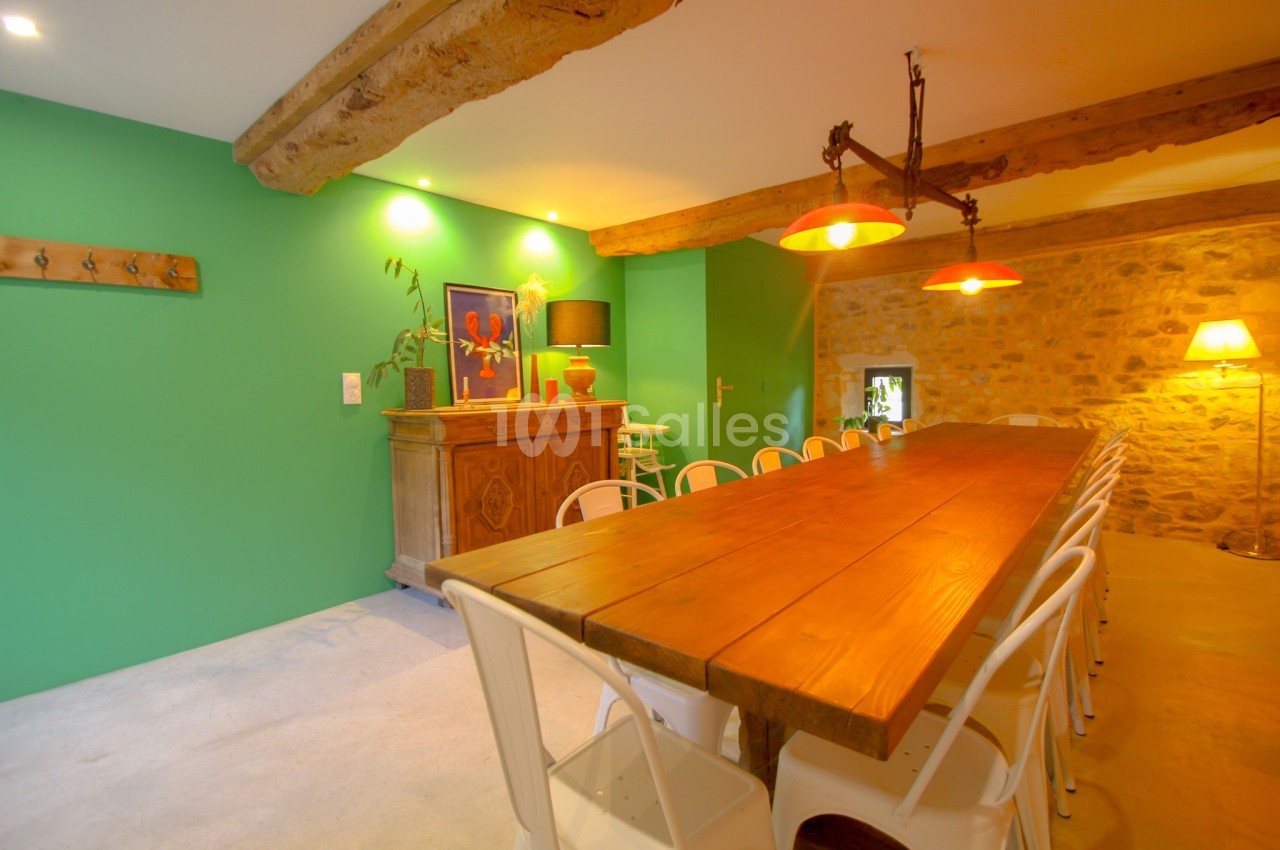 Salle à manger avec grande table en bois, chaises blanches, mur vert, poutres apparentes et éclairage chaleureux.