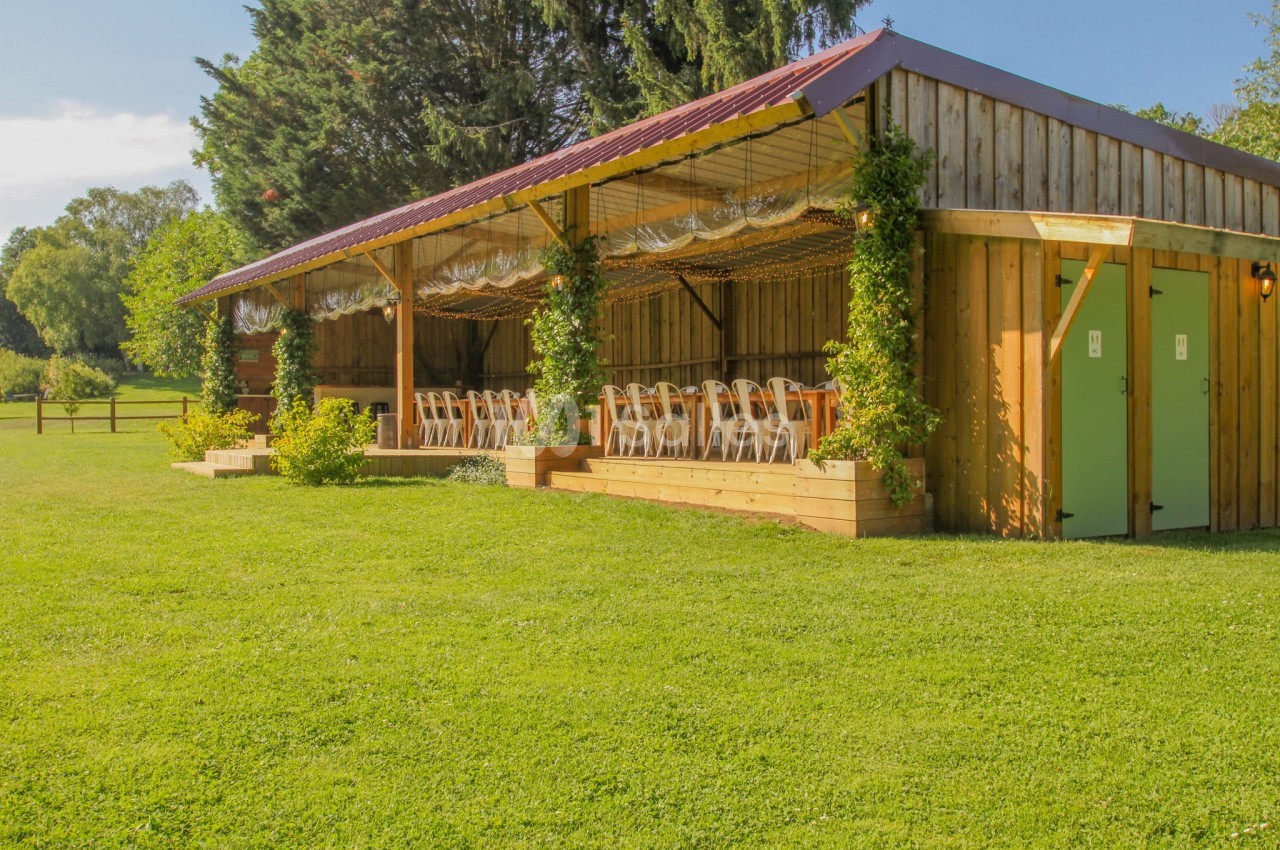Abri en bois avec des chaises alignées sous une pergola, entouré de verdure et éclairé par la lumière du jour.