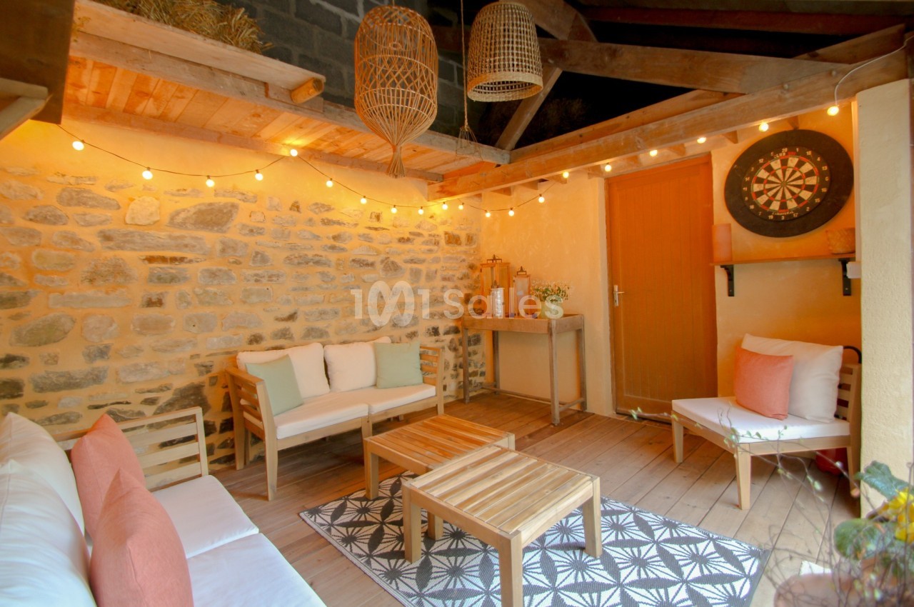 Espace extérieur couvert avec murs en pierre, mobilier en bois, guirlandes lumineuses et cible de fléchettes.
