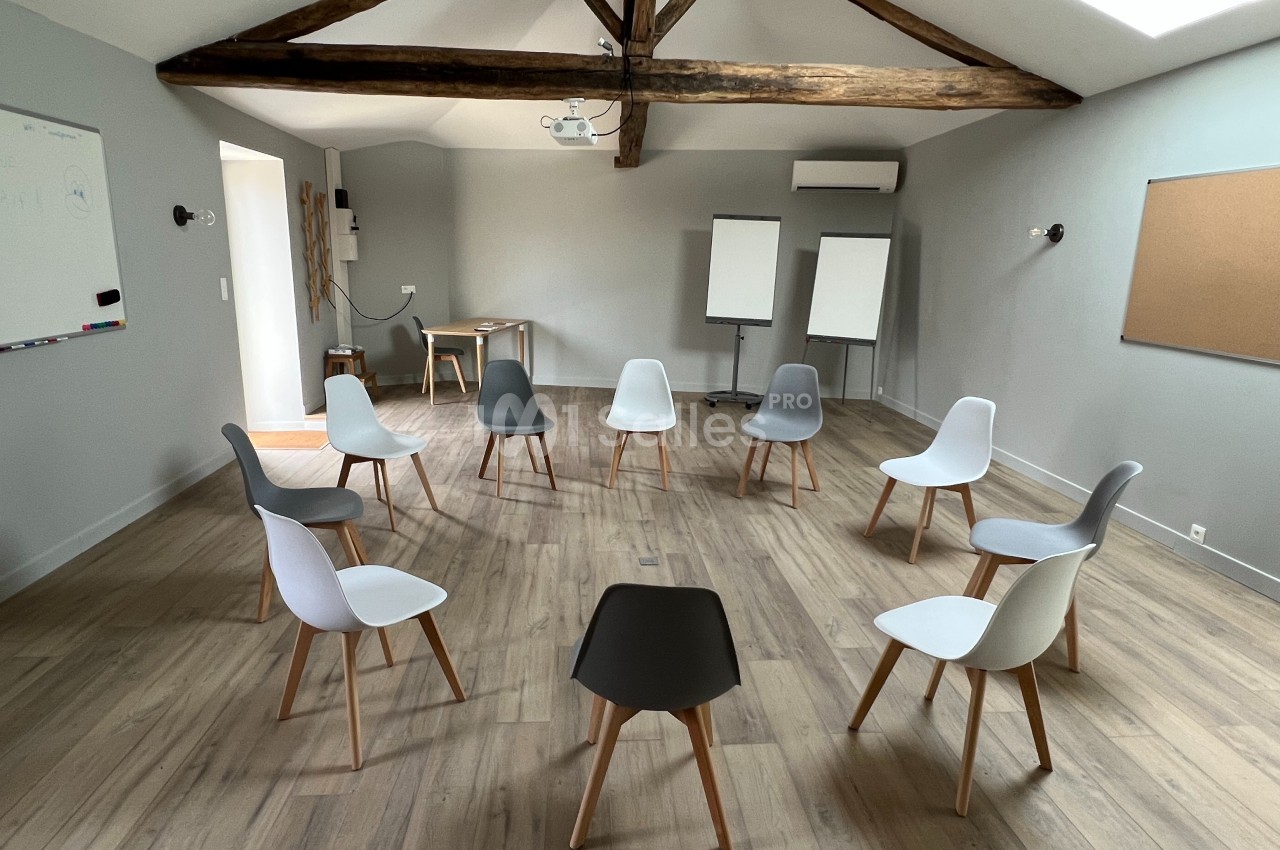 Salle lumineuse avec des chaises disposées en cercle, un tableau blanc, des chevalets et une table en arrière-plan.