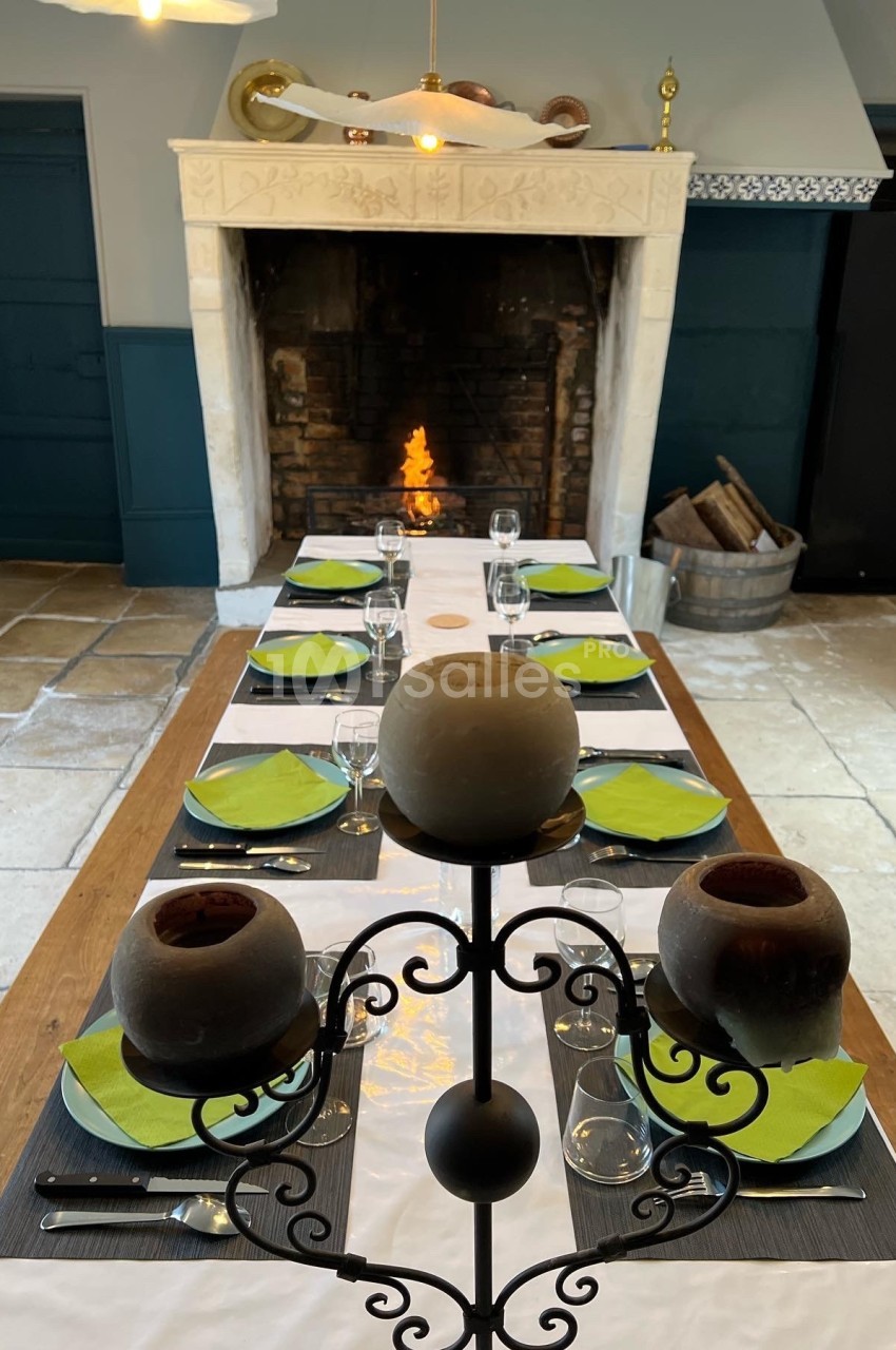 Table dressée avec des assiettes vertes et des verres, devant une cheminée allumée dans une pièce au sol en pierre.