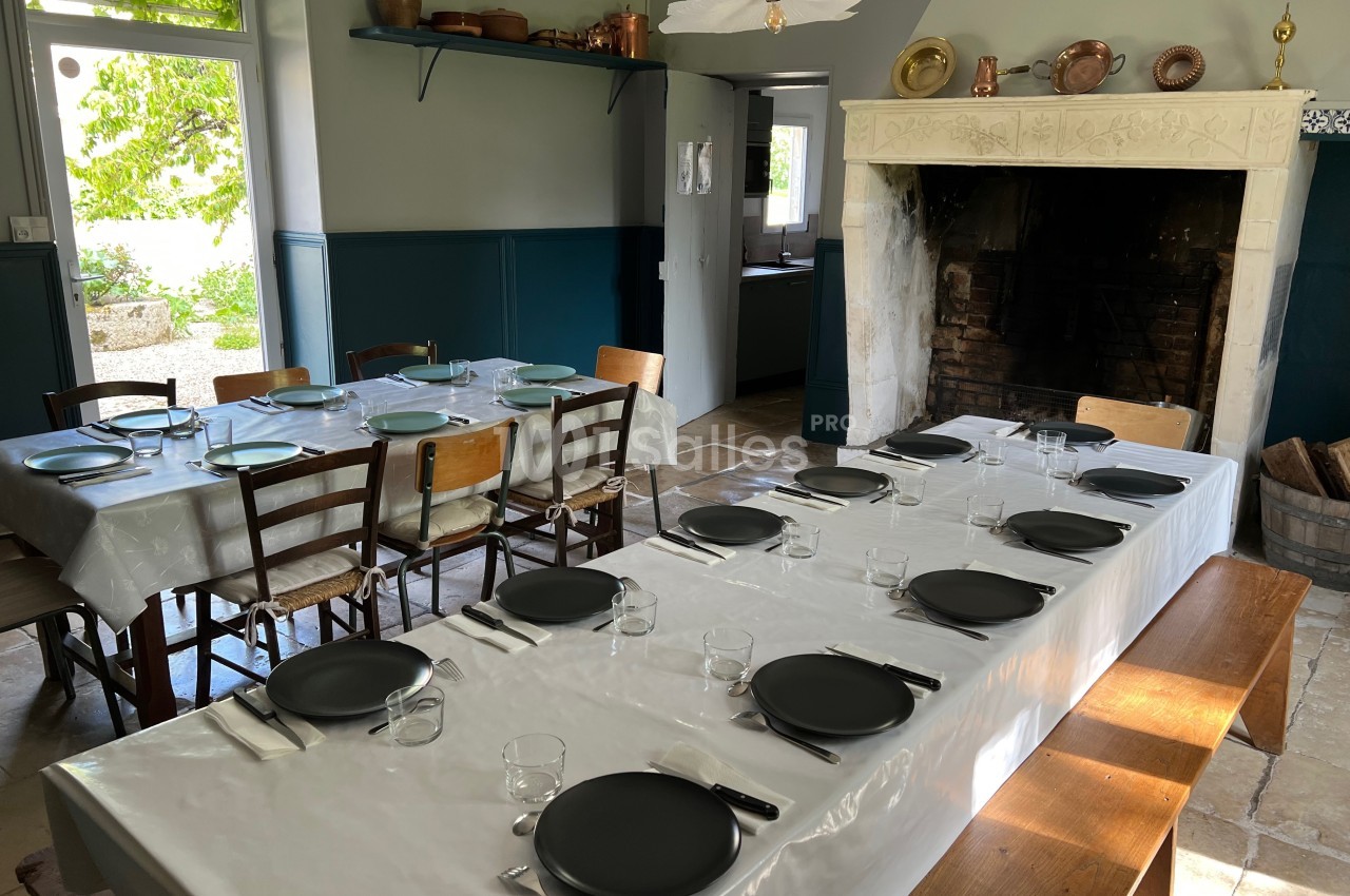 Grande table dressée avec des assiettes noires et des couverts dans une salle à manger rustique avec cheminée et lumière…