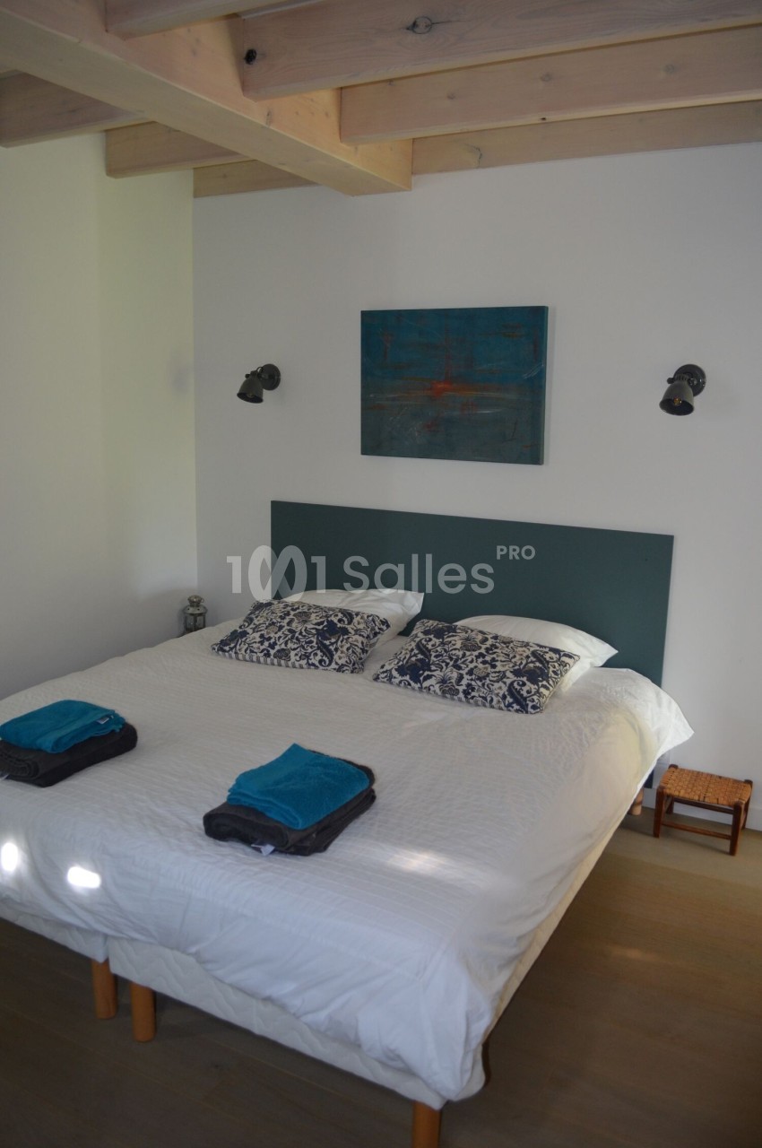 Lit double avec tête de lit verte, draps blancs, coussins à motifs, serviettes pliées et éclairage mural dans une chambre…