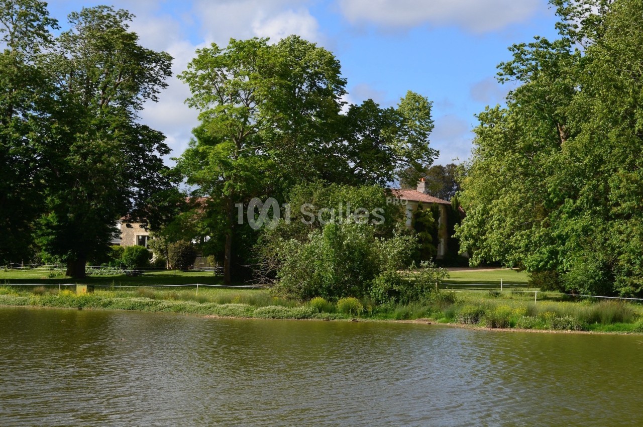 Vue d'un lac entouré de verdure avec des arbres et une maison en arrière-plan sous un ciel partiellement nuageux.