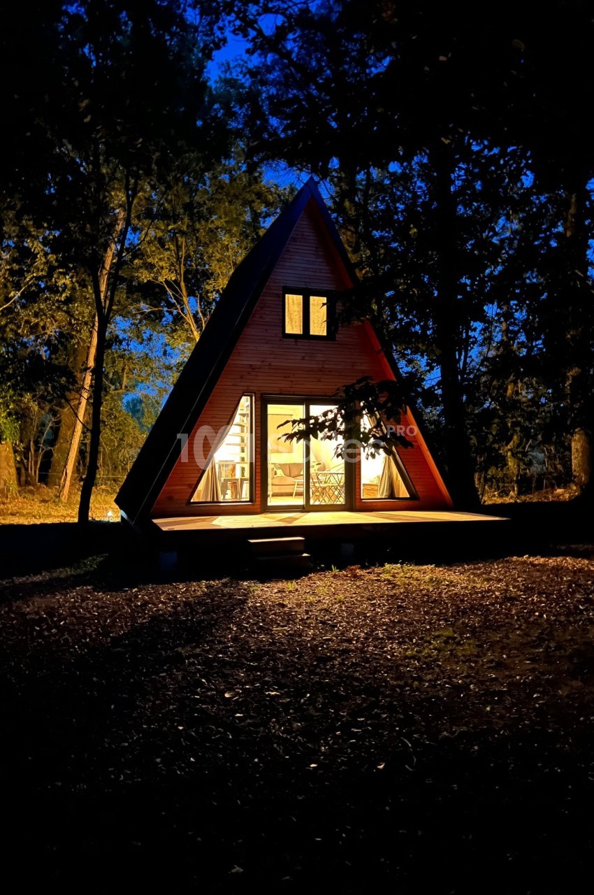 Cabane en forme de A éclairée de l'intérieur, entourée d'arbres dans un environnement nocturne.