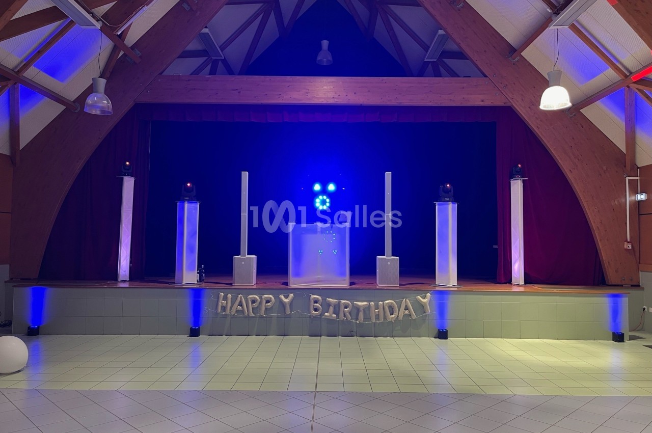 Salle décorée pour un anniversaire avec une scène illuminée en bleu, des ballons ’Happy Birthday’ et des équipements audio.