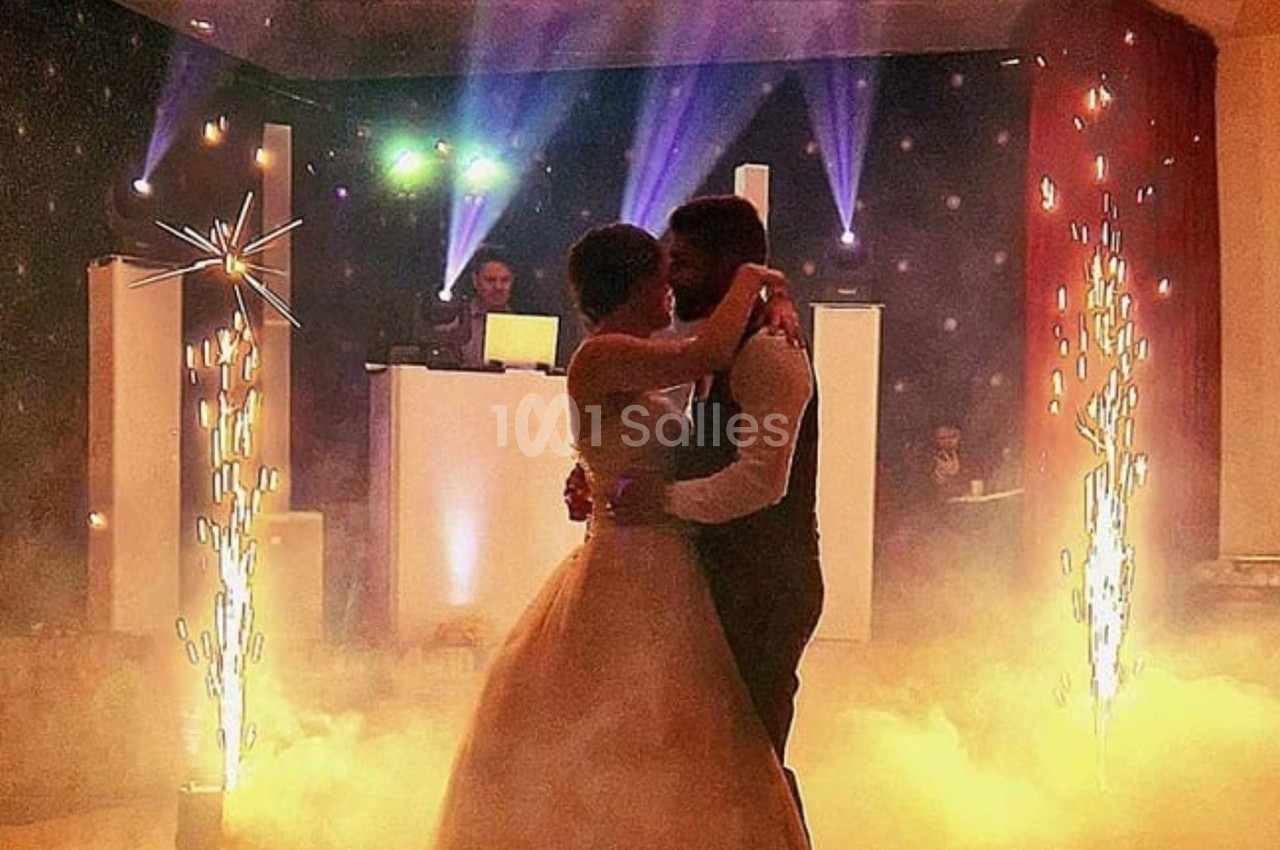 Un couple danse dans une ambiance festive avec éclairages colorés, fumée et étincelles, un DJ en arrière-plan.