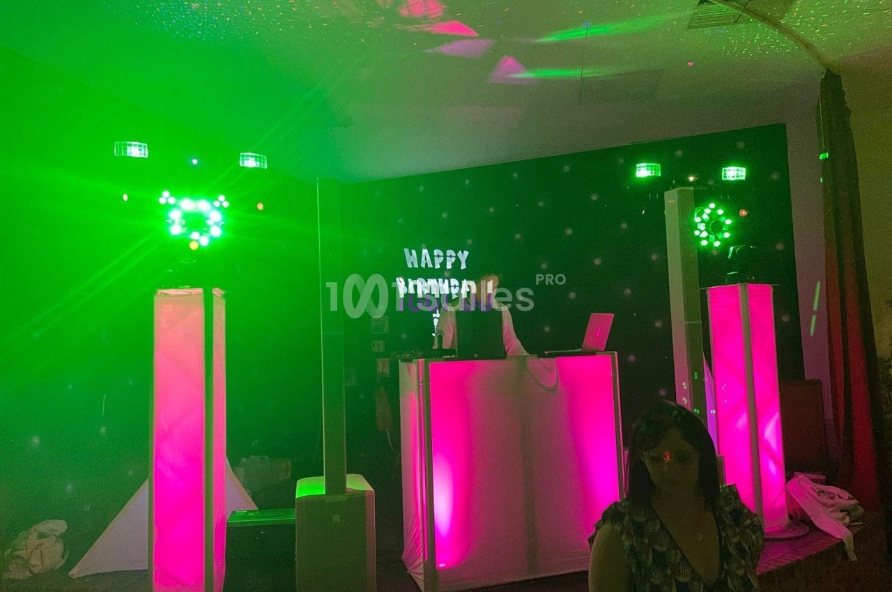 Un DJ anime une soirée avec des lumières vertes et roses sur une scène décorée d'un panneau ’Happy Birthday’.