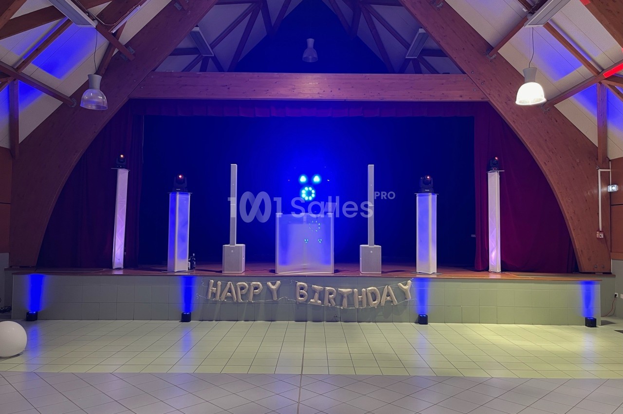 Salle décorée pour un anniversaire avec une scène illuminée en bleu, des ballons ’Happy Birthday’ et des équipements audio.