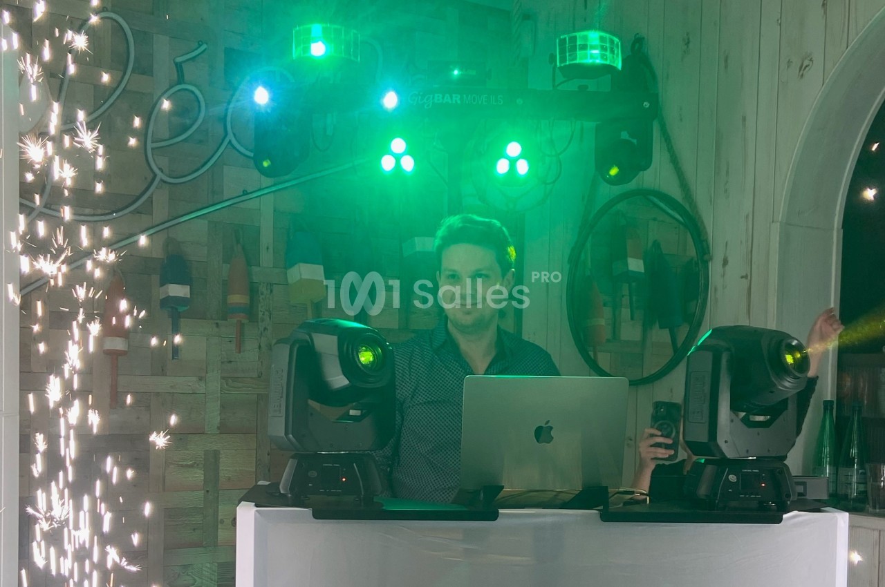 Un DJ mixe derrière une table équipée de projecteurs et d'un ordinateur, entouré d'effets lumineux et d'étincelles.