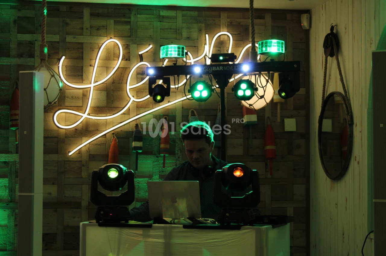 Un DJ mixe sur un ordinateur portable, entouré de lumières colorées dans un décor en bois avec des bouées suspendues.