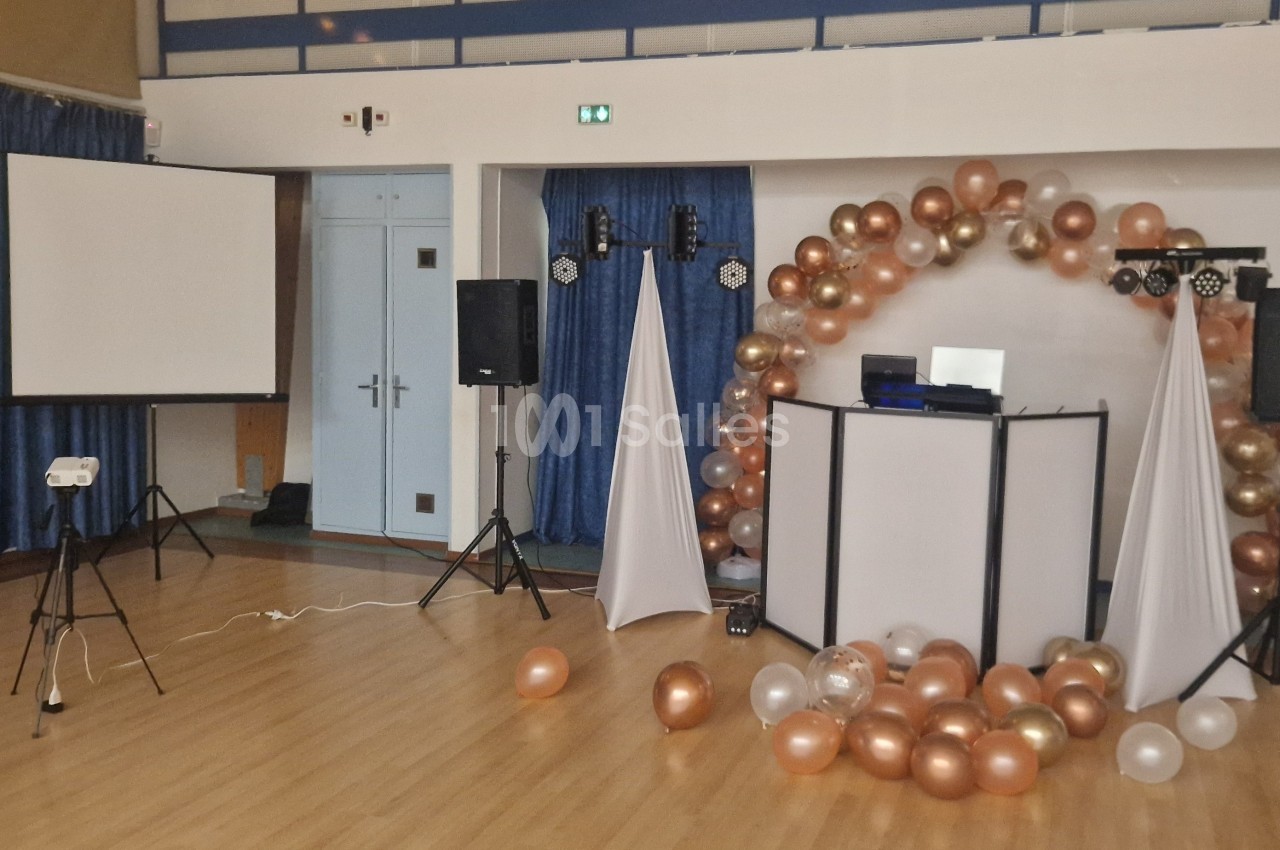 Salle décorée pour un événement avec arche de ballons dorés et blancs, projecteur, écran et matériel de sonorisation.