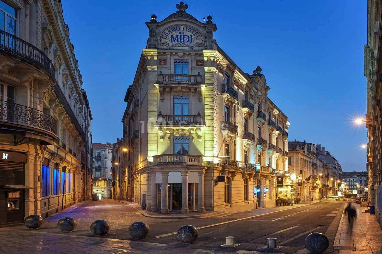 Façade éclairée du Grand Hôtel du Midi dans une rue calme au crépuscule, entourée de bâtiments historiques.