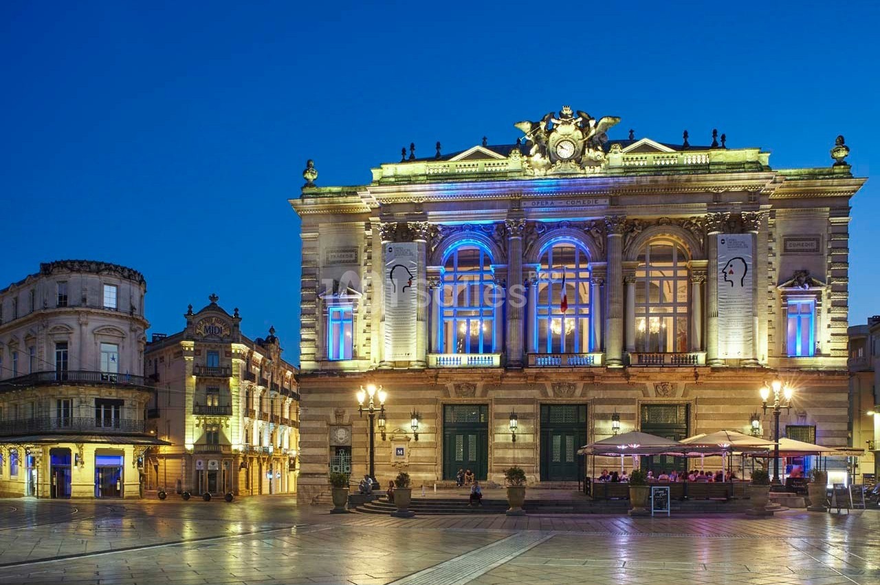 Façade illuminée d'un théâtre historique avec terrasse de café en soirée sur une place pavée.