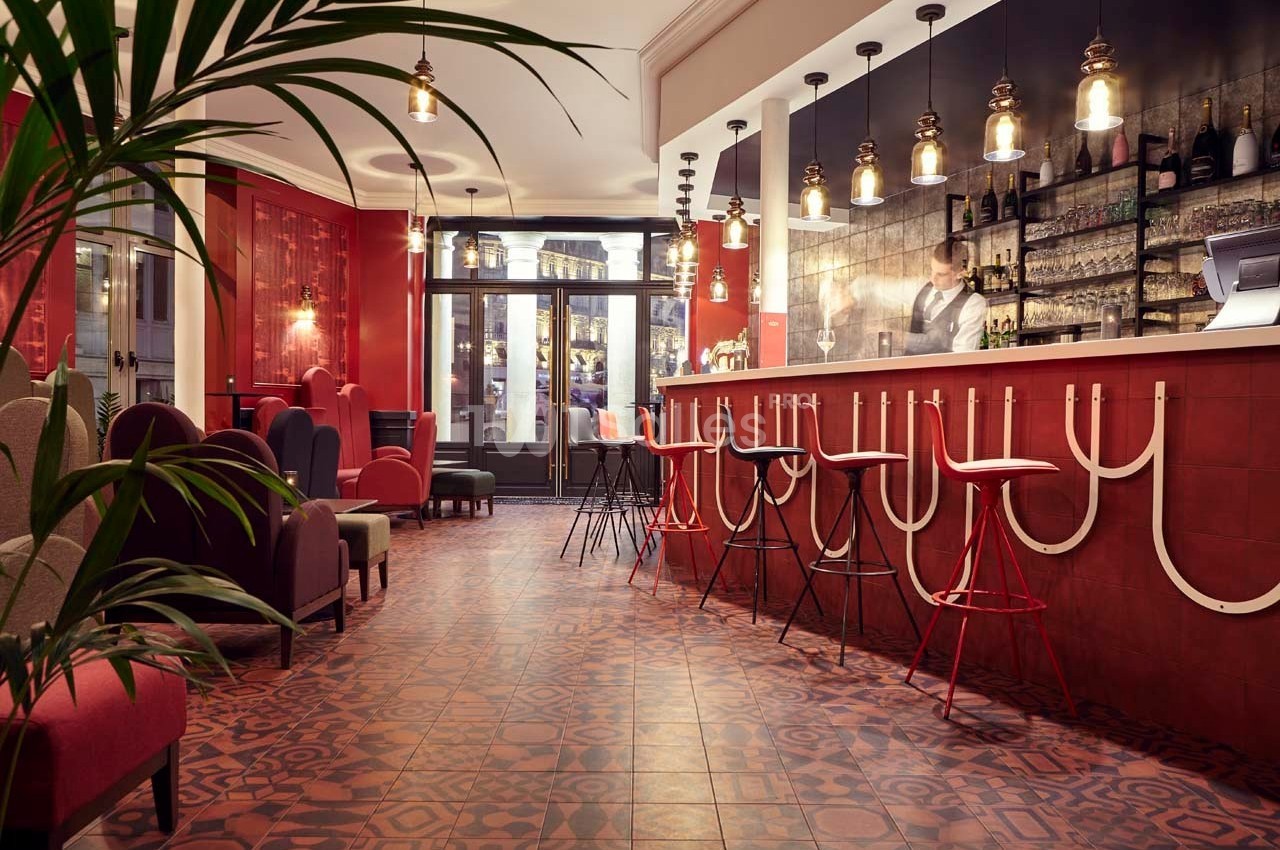 Intérieur d'un bar avec comptoir rouge, tabourets hauts, éclairage suspendu et fauteuils dans un décor chaleureux.