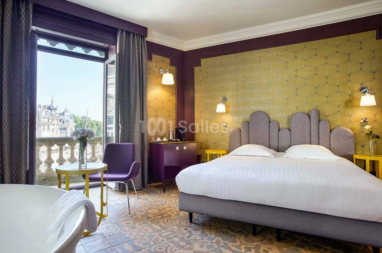 Chambre d'hôtel lumineuse avec grand lit, mobilier moderne, balcon donnant sur un paysage urbain et sol en carreaux…