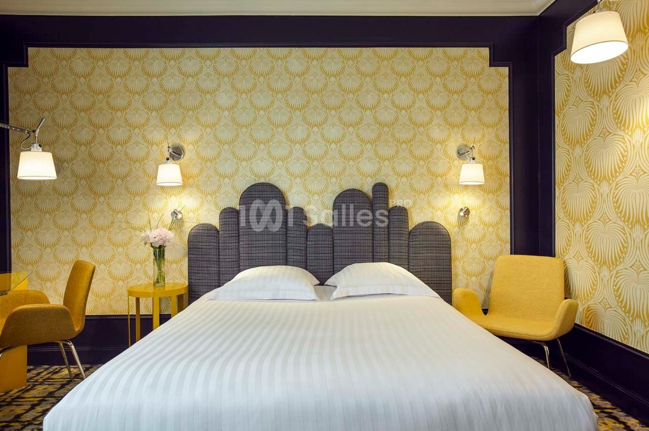 Chambre lumineuse avec lit double, tête de lit grise, fauteuils jaunes, lampes murales et papier peint à motifs dorés.