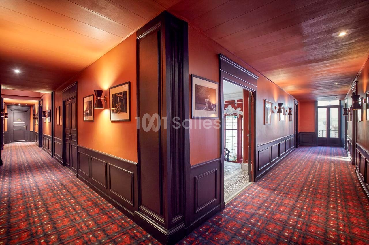 Couloir d'hôtel avec moquette rouge, murs orange, éclairage mural et portes menant à différentes pièces.