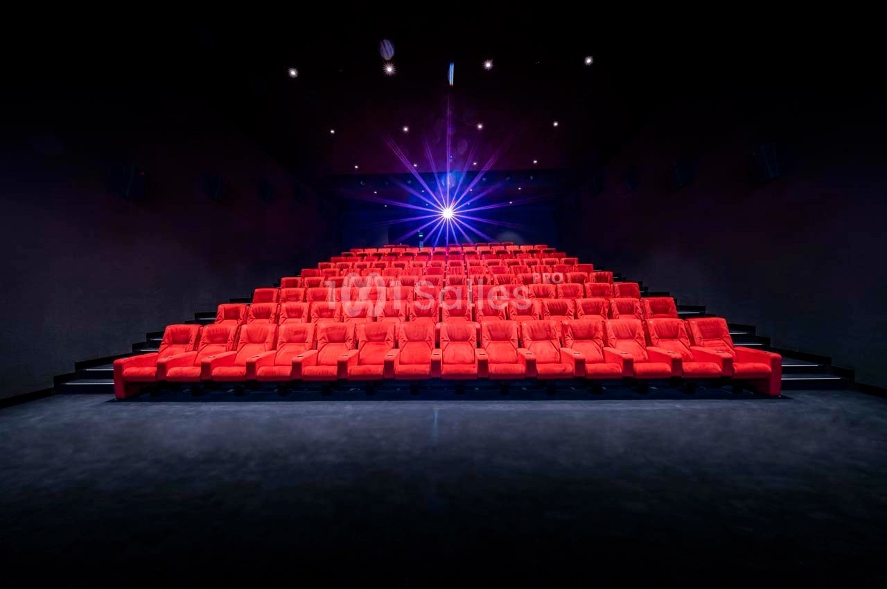 Salle de cinéma vide avec des rangées de fauteuils rouges et un projecteur allumé au fond.