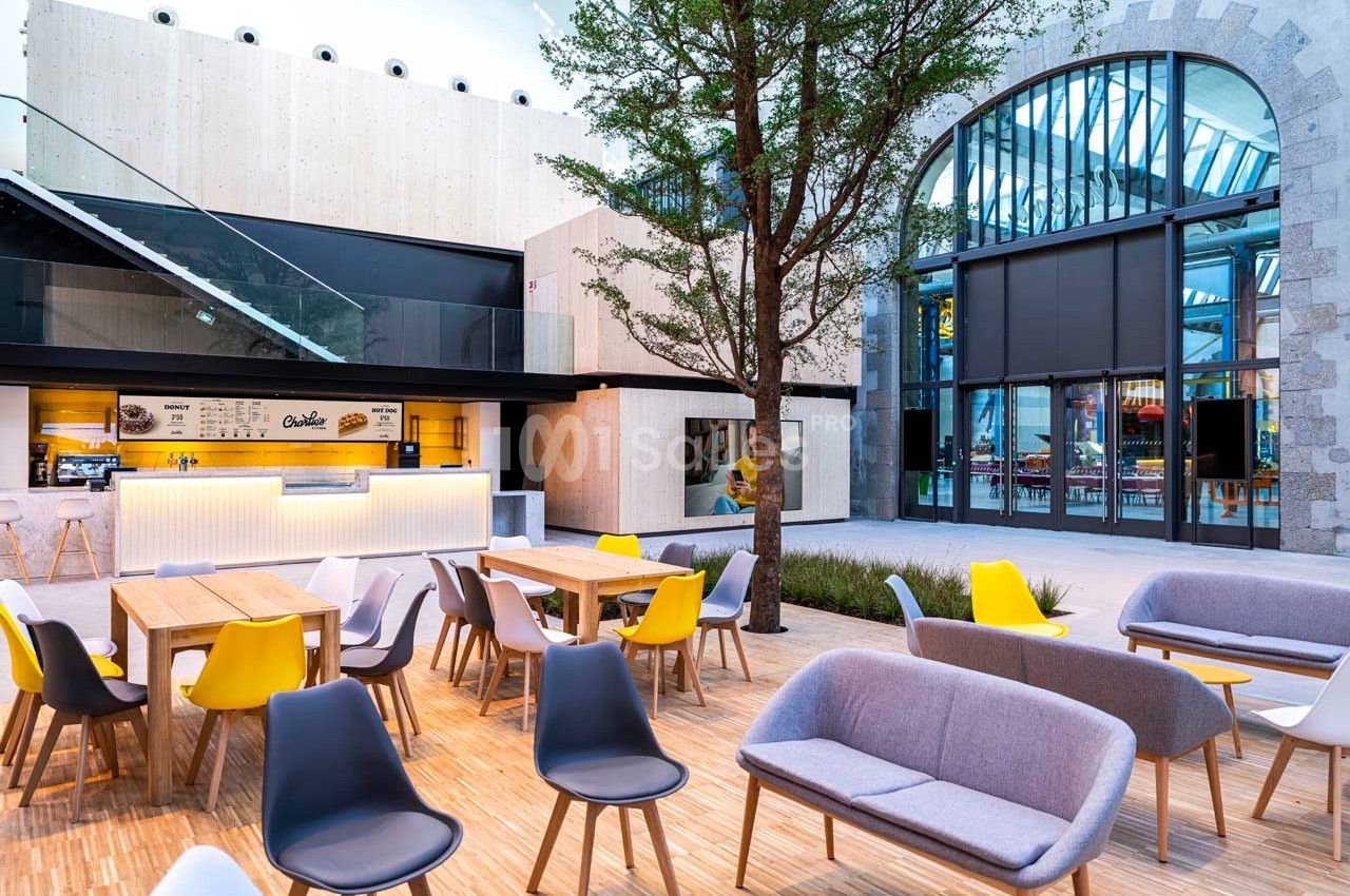 Terrasse moderne avec tables, chaises colorées et canapés, située devant un bâtiment vitré et un arbre central.