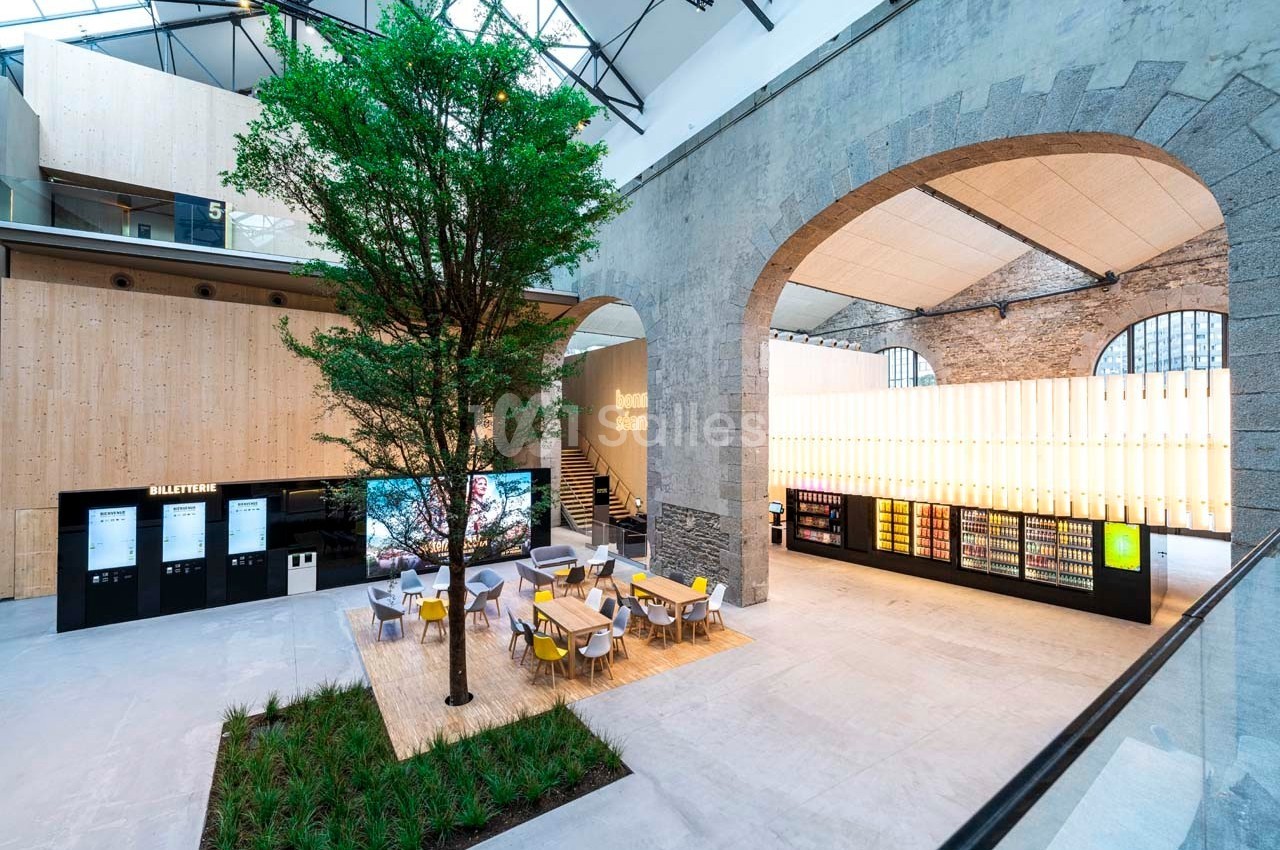 Espace intérieur moderne avec un arbre central, des tables, des distributeurs automatiques et des murs en pierre et bois.